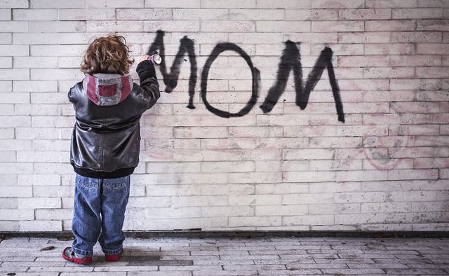 Small boy graffiti spraying 'mom' onto a white brick wall.