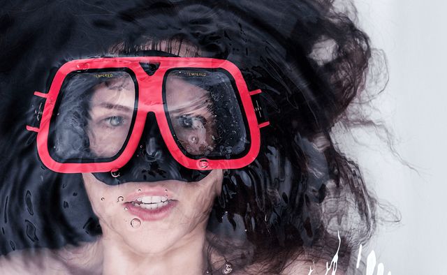 Woman's face with snorkelling goggles submerged in bath. Hair floats around her face. Tiny air bubbles escape her open mouth.