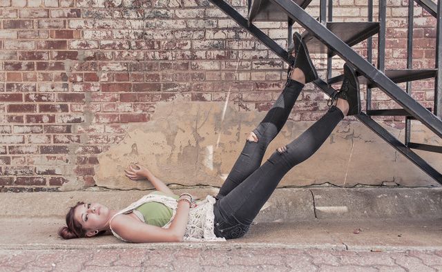 A woman lying on her back beside a brick wall. She has one hand on the brickwork and her feet resting on the edge of two steps of an open metal staircase above her.