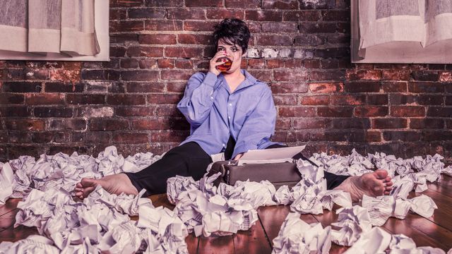 Woman with scruffy hair and makeup. On floor with typewriter in sea of screwed up paper. Blank stare, sipping whiskey glass.