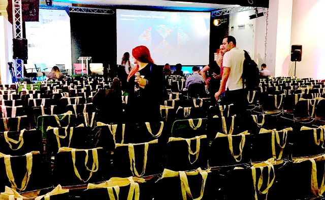 View of empty stage from back of room. People are starting to arrive to take their seat which are all draped with swag bags.